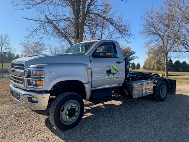 A silver flatbed pickup truck from a junk removal franchise parked on grass near trees on a sunny day.