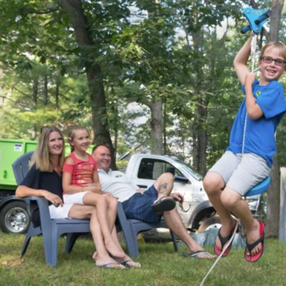 A boy rides a zipline in a backyard while a woman, a girl, and a man sit on chairs, smiling. A shed and a green truck are visible in the background.