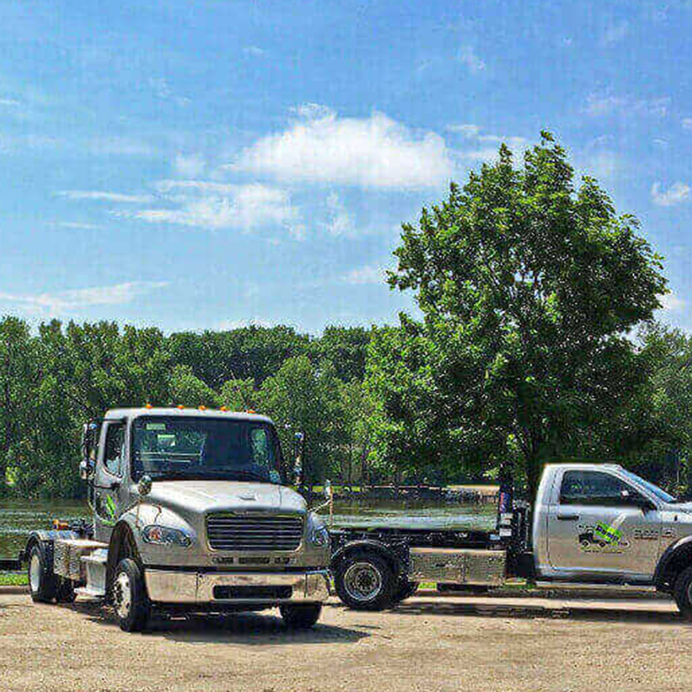 Three towing trucks parked in a row by a lake with trees, operating as part of a junk hauling franchise on a sunny day.